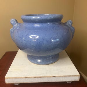May include: A blue ceramic vase with two side handles. The vase has a speckled finish and sits on a white wooden stand. The vase has a wide opening and a rounded body. The stand is rectangular and has small feet.