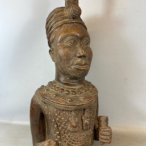 Puede incluir: Una escultura de madera marrón oscuro de una persona que lleva una corona y joyas elaboradas. La escultura está tallada con intrincados detalles y tiene un estilo africano tradicional.