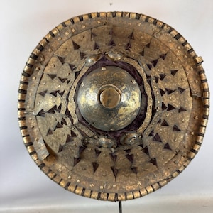 May include: A round, antique shield with a metallic gold finish. The shield features a central boss and is adorned with triangular and circular embellishments. The outer rim is bordered with a dark, textured material, and the shield is mounted on a black stand.