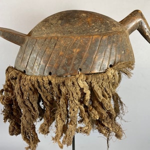 Puede incluir: Una máscara de madera con un pico largo y curvo y una franja de fibras marrones. La máscara está tallada con intrincados detalles y tiene un aspecto desgastado y antiguo.