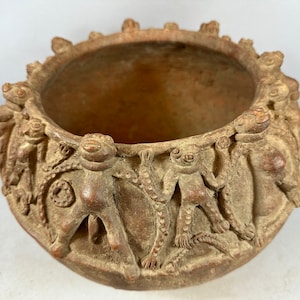 Op de afbeelding: Een terracotta pot met een ronde opening en een decoratief ontwerp van kikkers rond de rand. De kikkers staan in verschillende poses en hebben gedetailleerde kenmerken.
