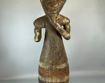 May include: A wooden sculpture of a person with a stylized head and body. The sculpture is painted in shades of brown, red, and white. The person is standing with their arms crossed and has a simple, geometric design.