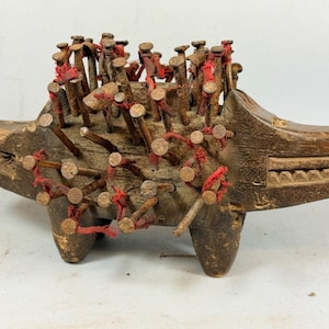 Puede incluir: Una figura animal de madera tallada con un hocico largo y cuatro patas, que presenta numerosos clavos de metal oxidados incrustados en el lomo y los costados. Un hilo rojo fino está enrollado alrededor de varios clavos, creando una textura.