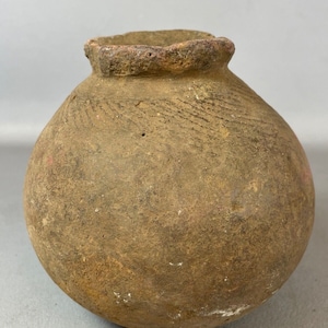 Può includere: Un vaso rotondo in terracotta marrone con un bordo leggermente svasato. Il vaso ha una superficie ruvida e testurizzata e sembra essere fatto a mano.