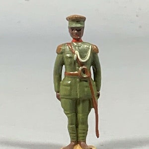 Puede incluir: Una figurita de soldado de juguete pintada de verde con un sombrero rojo y marrón, un cinturón blanco y una espada roja y marrón. El soldado está de pie sobre una base de color beige.