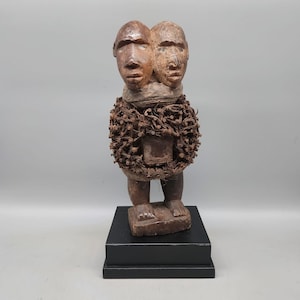 Puede incluir: Una escultura de madera tallada con dos cabezas unidas sobre un torso cubierto por una capa densa de clavos metálicos oxidados y material fibroso. La figura se asienta sobre una base rectangular y muestra una textura marrón desgastada.