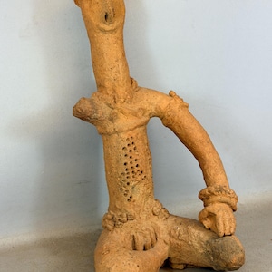 May include: A terracotta sculpture of a seated figure with an elongated neck and arms. The figure has a series of holes in the torso and is posed with one arm bent and resting on its knee. The sculpture is a light brown color.