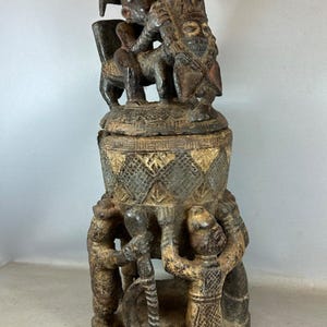 Könnte beinhalten: Geschnitzte Holzskulptur mit einer Figur, die auf einem Tier reitet, auf einem zylindrischen Sockel. Der Sockel ist mit geschnitzten Figuren und geometrischen Mustern verziert. Die Skulptur ist hauptsächlich braun mit helleren Akzenten.