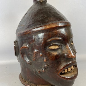 Può includere: Una scultura in legno marrone scuro di una testa umana con la parte superiore arrotondata. La testa ha un viso scolpito con una bocca larga e denti bianchi. La scultura è un'opera d'arte africana tradizionale.