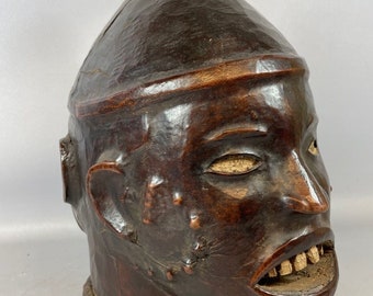 May include: A dark brown wooden sculpture of a human head with a rounded top. The head has a carved face with a wide mouth and white teeth. The sculpture is a traditional African art piece.