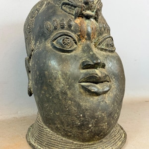 Puede incluir: Una escultura de bronce de una cabeza humana con un rostro detallado y una figura de león en la parte superior de la cabeza. La escultura tiene una pátina oscura y envejecida y un collar detallado. Los ojos están abiertos y los labios ligeramente entreabiertos.