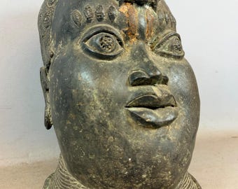 Pode incluir: Uma escultura em bronze de uma cabeça humana com um rosto detalhado e uma figura semelhante a um leão no topo da cabeça. A escultura tem uma pátina escura e envelhecida e uma gola detalhada. Os olhos estão abertos e os lábios ligeiramente entreabertos.