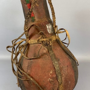 Könnte beinhalten: Eine braune Lederflasche mit einem geflochtenen Ledergriff und einem Perlenmuster. Die Flasche ist mit Lederriemen verziert und hat eine dunkelbraune Oberfläche.