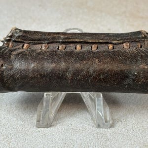 Könnte beinhalten: Zylindrisches, dunkelbraunes Lederobjekt, möglicherweise ein Behälter oder eine Tasche, mit leicht abgenutztem Aussehen. Das Leder ist genäht und hat kleine, dekorative Holzstifte entlang der Oberseite. Es ruht auf einem transparenten, abgewinkelten Präsentationsständer.