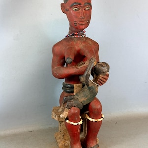 Può includere: Statua in legno scolpito di una figura seduta con la pelle rossa, che indossa una corona scura e una collana di perline. La figura tiene in braccio un bambino dalla pelle scura. La statua è dipinta con accenti rossi, neri e bianchi ed è seduta su uno sgabello.