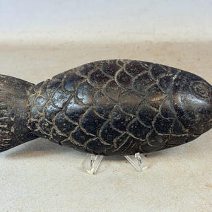 Peut inclure: Sculpture de poisson sculptée en marron foncé, avec des motifs d'écailles détaillés. Le poisson a une nageoire caudale proéminente et un œil détaillé. La sculpture est présentée sur un support transparent.
