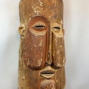 以下が含まれることがあります： 長方形の形をした、風化した木製のマスク。様式化された人間の顔が特徴で、細長い目の穴、突き出た鼻、小さな開いた口があります。色は主に薄茶色で、赤茶色のアクセントがあります。