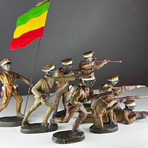 Può includere: Un gruppo di soldatini giocattolo vintage in uniforme kaki, alcuni puntano fucili. Un soldato tiene una bandiera a strisce rosse, gialle e verdi. Le figure sono su una superficie bianca, sfondo grigio.