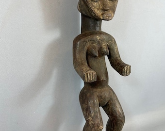 May include: A dark brown wooden sculpture of a standing female figure. The figure has a stylized head with a crown and a simplified body with exaggerated features.