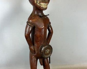 May include: A carved wooden figure with a dark brown finish. The figure has a stylized face with inlaid eyes and a wide grin. Metal elements are affixed to the head and body. The figure stands on a rectangular base, and holds a circular object.