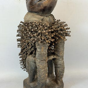 Peut inclure: Une figurine en bois sculpté brun foncé avec une tête lisse et arrondie sur une base. Le torse est densément recouvert de nombreux clous métalliques saillants et enveloppé dans une cordelette en fibre épaisse, texturée et usée.