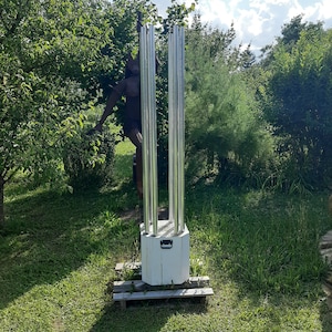 May include: An outdoor light fixture with multiple vertical, silver tubes. The fixture is mounted on a white, wooden base and sits on a wooden platform. The background features green foliage and a statue.