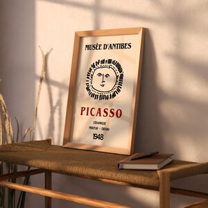 May include: A framed art print with a Picasso design. The print has a beige background with the words "Mus&eacute;e d'Antibes" at the top and "Picasso" in red. Below is a sun-like illustration and the year 1948.
