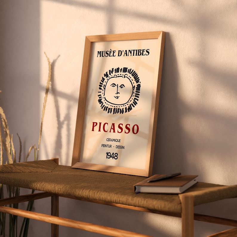 May include: A framed art print with a Picasso design. The print has a beige background with the words "Mus&eacute;e d'Antibes" at the top and "Picasso" in red. Below is a sun-like illustration and the year 1948.