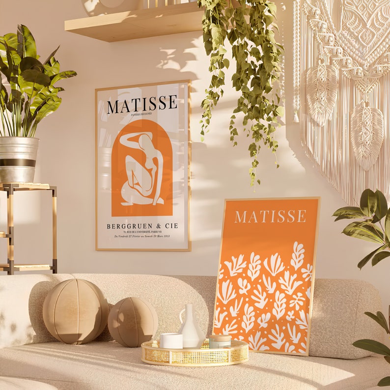 Puede incluir: Dos impresiones enmarcadas con el nombre "Matisse" en texto blanco sobre fondo naranja. La impresi&oacute;n de la izquierda presenta una silueta blanca de una mujer sentada con la cabeza apoyada en la mano. La impresi&oacute;n de la derecha presenta un dise&ntilde;o abstracto blanco de hojas y ramas.