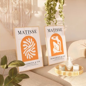 Puede incluir: Dos impresiones de arte enmarcadas con fondo blanco y acentos naranjas. Las impresiones presentan el texto "MATISSE" y el nombre de una galer&iacute;a. Una impresi&oacute;n presenta una forma naranja abstracta y la otra presenta una figura desnuda en posici&oacute;n sentada.