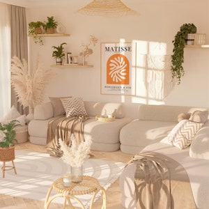 Puede incluir: Una sala de estar con un sof&aacute; seccional beige, una alfombra blanca y una mesa auxiliar con un jarr&oacute;n de hierba de pampa seca. La pared detr&aacute;s del sof&aacute; presenta una impresi&oacute;n enmarcada de un dise&ntilde;o abstracto naranja y blanco con el texto "MATISSE" y "BERGGRUEN & CIE".