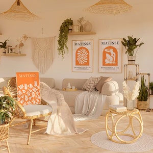 Puede incluir: Tres impresiones de arte Matisse naranja y blanco con el texto "MATISSE" y "BERGGRUEN & CIE" en una sala de estar con un sof&aacute; beige, una silla de mimbre, una mesa auxiliar de mimbre y plantas.