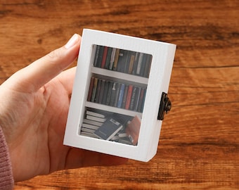 May include: White miniature bookcase with glass doors and a small collection of books inside. The bookcase is closed with a small metal latch.