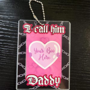 May include: A clear acrylic tag with a silver chain. The tag features the text "I call him Daddy" and "Your Boo Here" in a pink heart design. A chain pattern borders the pink background.