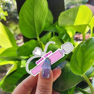 May include: A pink and white sticker with a glitter design of a toy with the text "Good Vibes" on it.