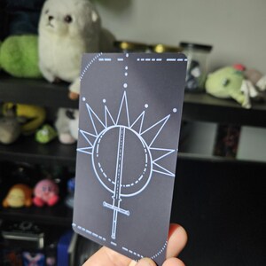 May include: A rectangular playing card with a dark gray background featuring a white line art design of a sword pointing downward, centered within a circle and surrounded by a radiating starburst pattern with small dots at the tips of the lines.