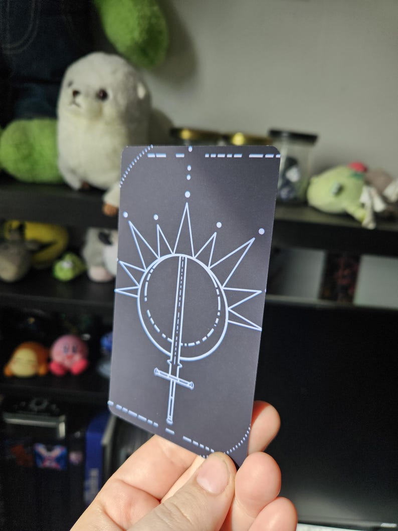 May include: A rectangular playing card with a dark gray background featuring a white line art design of a sword pointing downward, centered within a circle and surrounded by a radiating starburst pattern with small dots at the tips of the lines.