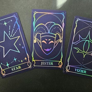 May include: Three rectangular tarot cards with a dark blue background and holographic foil outlines. The left card features a star and the text STAR. The center card displays a jester mask and the text JESTER. The right card shows an eye and the text VIZIER.