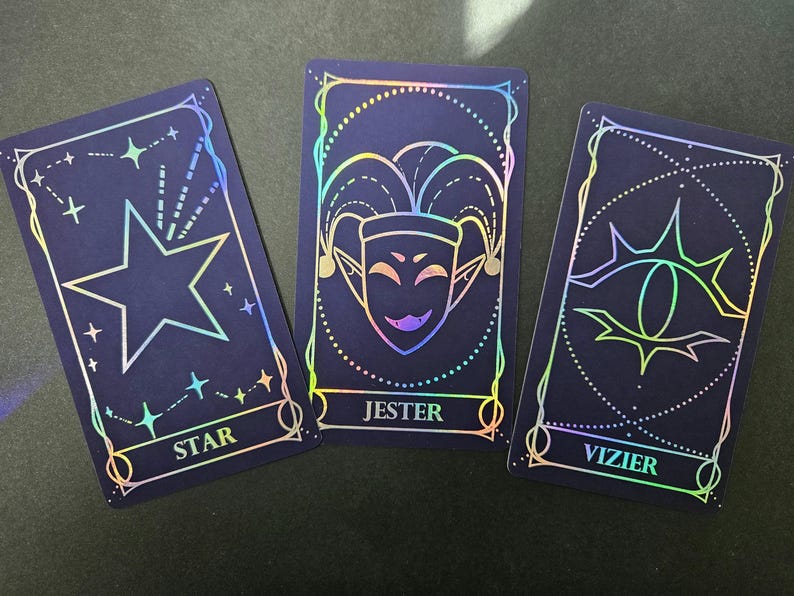 May include: Three rectangular tarot cards with a dark blue background and holographic foil outlines. The left card features a star and the text STAR. The center card displays a jester mask and the text JESTER. The right card shows an eye and the text VIZIER.