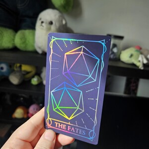 May include: A rectangular card with a dark purple background featuring two holographic 20-sided dice outlines. The card has a decorative border and the text THE FATES printed in a holographic font at the bottom center of the design.