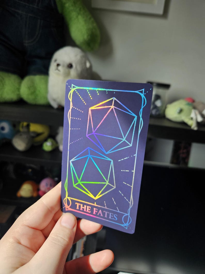 May include: A rectangular card with a dark purple background featuring two holographic 20-sided dice outlines. The card has a decorative border and the text THE FATES printed in a holographic font at the bottom center of the design.