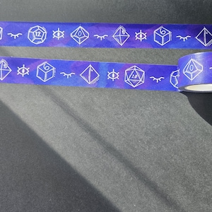 May include: A roll of washi tape with a purple and blue watercolor background. The tape features white outlines of dice and eyes. The tape is on a dark gray surface.