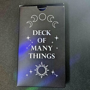 May include: A rectangular black card deck box featuring the white text DECK OF MANY THINGS. The design includes a lunar cycle graphic at the top, a sun icon at the bottom, and small star symbols scattered across the front surface with blue highlights.