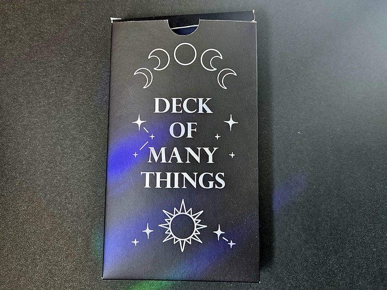 May include: A rectangular black card deck box featuring the white text DECK OF MANY THINGS. The design includes a lunar cycle graphic at the top, a sun icon at the bottom, and small star symbols scattered across the front surface with blue highlights.