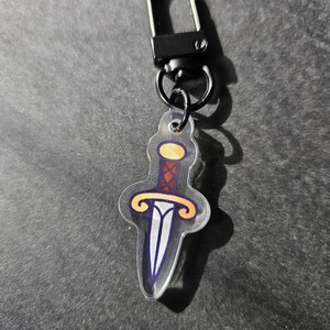 May include: A clear acrylic keychain with a cartoon dagger design. The dagger is blue, white, and yellow with a brown handle.