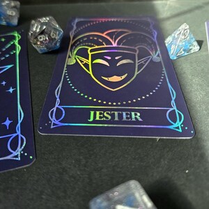 May include: A dark blue playing card featuring a holographic jester face icon and the text JESTER in a bold font. The card is surrounded by several multi-faceted, translucent blue and silver dice with visible numbers, creating a gaming aesthetic.