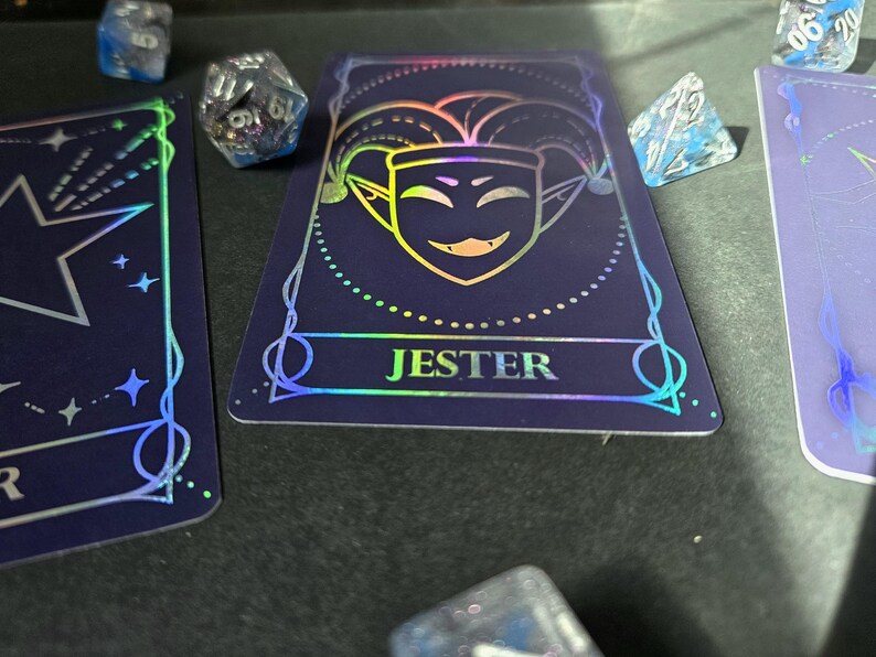 May include: A dark blue playing card featuring a holographic jester face icon and the text JESTER in a bold font. The card is surrounded by several multi-faceted, translucent blue and silver dice with visible numbers, creating a gaming aesthetic.