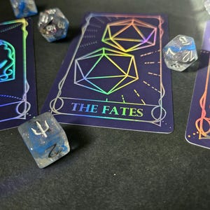 Könnte beinhalten: Ein Set aus polyedrischen Würfeln und Tarotkarten mit holografischen Folienmotiven. Die mittlere Karte zeigt zwei Ikosaeder und den Text THE FATES. Weitere Karten zeigen Mondphasen und das Wort FOOL. Die Würfel sind durchscheinend blau und klar.