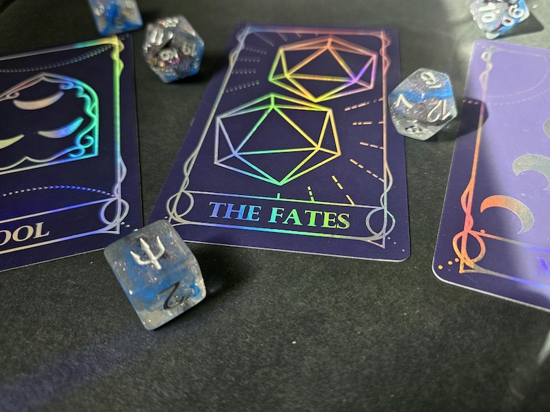 May include: A set of polyhedral dice and tarot cards featuring holographic foil designs. The central card displays two icosahedrons and the text THE FATES. Other cards show moon phases and the word FOOL. The dice are translucent blue and clear.