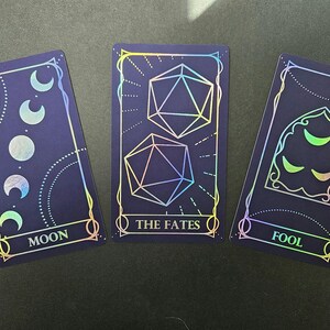 May include: Three rectangular cards with a dark blue background and holographic foil details. The first card shows moon phases and the text MOON. The second card features two icosahedron dice and the text THE FATES. The third card displays a gothic arch with crescent moons and the text FOOL.